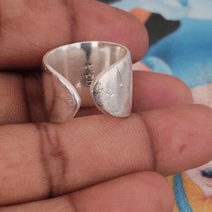 Può includere: Un anello aperto in argento a fascia larga con una superficie lucida e leggermente testurizzata. L'interno della fascia presenta un chiaro marchio 925, che indica una composizione in argento sterling. Il design è regolabile con due estremità arrotondate.