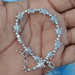 Puede incluir: Una pulsera de tono plateado que presenta una serie de pequeños dijes detallados de tortuga que se alternan con cuentas redondas y lisas de tono plateado. La pulsera incluye una cadena extensora de metal con un cierre de mosquetón ajustable.