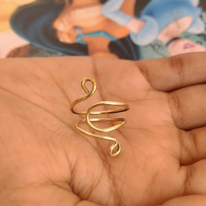 May include: A gold-coloured, wire-wrapped ring with a unique, swirling design. The ring features a spiral shape with two loops and a wrapped band, creating an elegant and artistic look. The ring is shown on a person's hand.