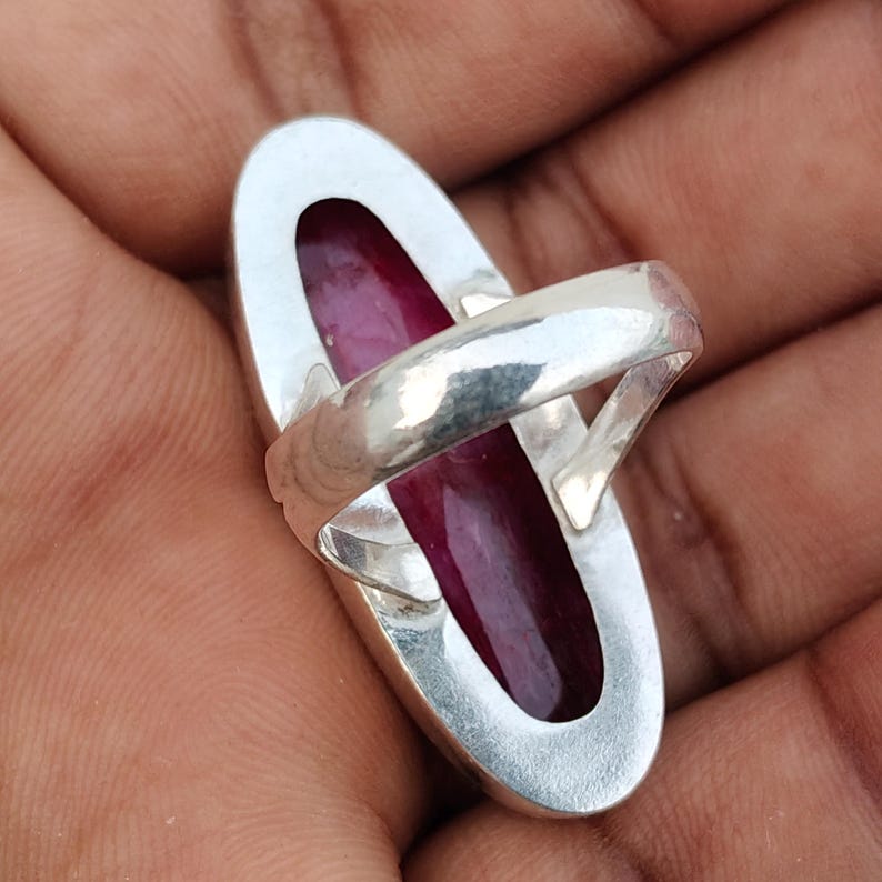 May include: A silver ring featuring a long, oval-shaped, deep red gemstone set within a polished metal bezel. The band is smooth and metallic, showcasing a simple, clean design that highlights the contrast between the dark red stone and silver.