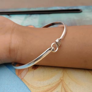 May include: A silver-colored bangle bracelet with a hook and eye closure. The bracelet is a simple, elegant design, with a smooth, polished finish. The closure is made of coiled wire and a small loop.