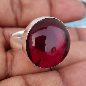 Könnte beinhalten: Ein Silberring mit einem großen, runden, polierten tiefroten Edelstein, der in einer glatten Metallfassung sitzt. Der Stein hat eine glänzende, reflektierende Oberfläche mit dunklen Einschlüssen im durchscheinenden, dunkelroten Material.