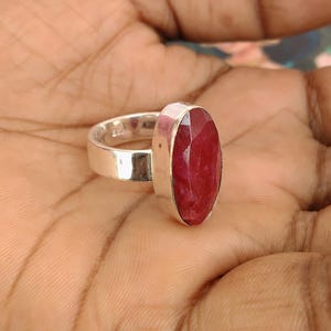 May include: A silver ring with a large, oval-cut ruby gemstone. The ruby is a deep red color and has many facets. The ring band is wide and made of polished silver. The ring is a piece of jewelry.