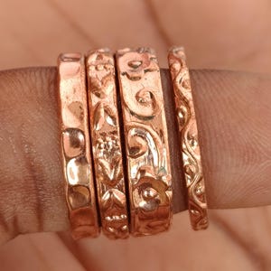 Puede incluir: Cuatro anillos de color cobre con intrincados diseños en relieve. Los anillos presentan patrones de puntos, motivos florales y líneas onduladas. Los anillos están apilados, mostrando la artesanía detallada y las texturas variadas.