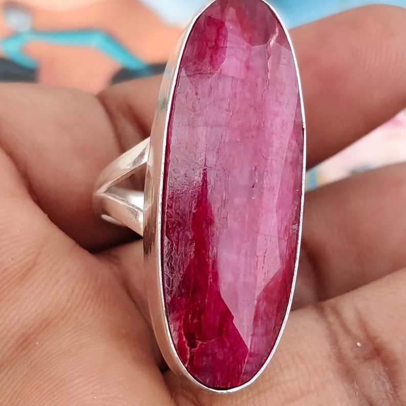 May include: A silver ring featuring a large, elongated oval-shaped faceted ruby gemstone. The deep pinkish-red stone is set in a smooth silver bezel with a split shank band design, showcasing the natural texture and facets of the gemstone.