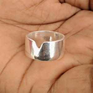 Könnte beinhalten: Ein silberner Ring mit einem einzigartigen V-förmigen Ausschnitt. Der Ring hat ein dickes Band und eine polierte Oberfläche, die das Licht reflektiert. Der Ring wird vor einem hautfarbenen Hintergrund gezeigt.