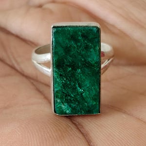 Peut inclure: Une bague en argent avec une grande pierre rectangulaire vert émeraude à la coupe brute, sertie dans une monture en métal lisse. L'anneau de la bague est divisé en deux fins segments d'argent qui rejoignent le sertissage central, révélant une surface cristalline texturée.