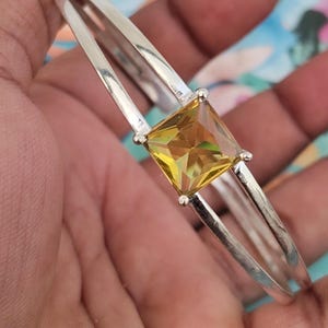 Puede incluir: Un brazalete de metal en tono plateado con un diseño de vástago dividido y una única piedra preciosa amarilla grande de corte cuadrado, engastada en el centro con cuatro garras. El brazalete tiene un acabado pulido y reflectante con una piedra amarilla facetada.