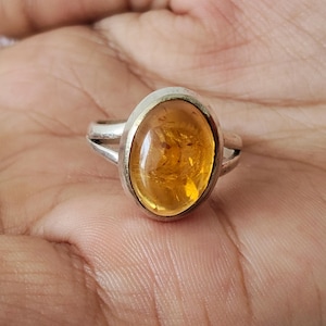 May include: A silver ring featuring a large, oval-shaped, translucent amber gemstone with a warm golden-orange hue. The stone is set in a smooth metal bezel, and the band consists of a split-shank design that tapers toward the back of the ring.