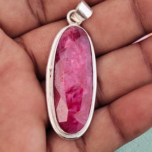May include: An elongated oval pendant featuring a faceted, deep pinkish-red gemstone set within a smooth, polished silver-toned metal frame. The gemstone displays a textured, multi-faceted surface, and the pendant includes a silver-toned bail at the top.
