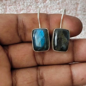 May include: A pair of rectangular drop earrings featuring polished labradorite gemstones set in silver-toned metal. One stone displays a vibrant blue flash, while the other shows a darker, muted grey tone with subtle iridescent reflections.