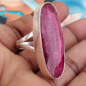 May include: A silver ring featuring a large, elongated oval-shaped faceted ruby gemstone set in a smooth metal bezel. The band of the ring has a split design, with two separate silver arms connecting to the main setting of the deep pink stone.