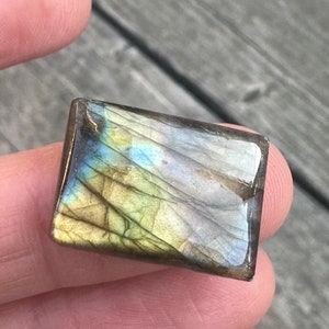 May include: A rectangular labradorite gemstone with iridescent blue, green, and yellow hues. The polished stone has a glossy finish and displays natural veining patterns. The gemstone is held between fingers.