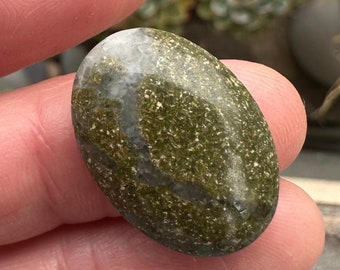 May include: An oval-shaped, polished stone with a speckled green and white pattern. The stone is held in a hand, showcasing its smooth texture and natural variations. The stone's surface has a mix of light and dark green hues.