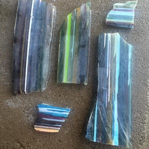 May include: Five pieces of broken glass with vertical stripes in shades of blue, green, white, and purple. The glass fragments vary in size and shape, with some having curved edges. The pieces are arranged on a rough, gray surface.