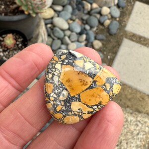 Puede incluir: Una piedra triangular con un patr&oacute;n de mosaico de fragmentos beige, negros y blancos. La piedra se sostiene en una mano, mostrando su superficie pulida y sus variaciones de color &uacute;nicas. El dise&ntilde;o de la piedra recuerda una est&eacute;tica natural y terrosa.