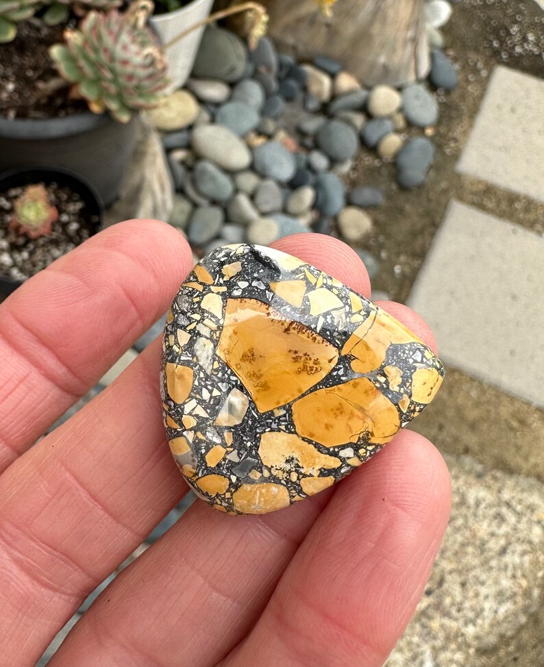 Puede incluir: Una piedra triangular con un patr&oacute;n de mosaico de fragmentos beige, negros y blancos. La piedra se sostiene en una mano, mostrando su superficie pulida y sus variaciones de color &uacute;nicas. El dise&ntilde;o de la piedra recuerda una est&eacute;tica natural y terrosa.
