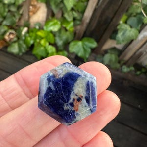 K&ouml;nnte beinhalten: Ein sechseckiger Edelstein mit tiefblauer Farbe, durchsetzt mit helleren grauen und braunen Flecken. Der polierte Stein wird in einer Hand gehalten, mit einem verschwommenen gr&uuml;nen Hintergrund aus Bl&auml;ttern und einem Holzzaun.