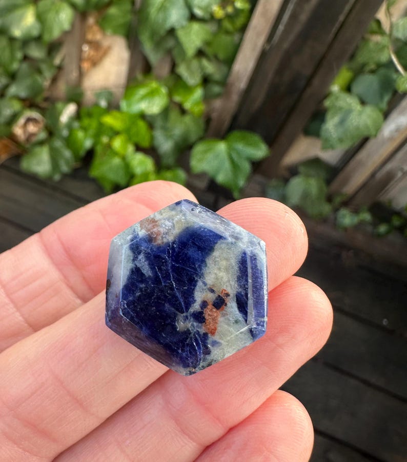 K&ouml;nnte beinhalten: Ein sechseckiger Edelstein mit tiefblauer Farbe, durchsetzt mit helleren grauen und braunen Flecken. Der polierte Stein wird in einer Hand gehalten, mit einem verschwommenen gr&uuml;nen Hintergrund aus Bl&auml;ttern und einem Holzzaun.