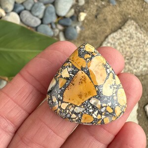 Puede incluir: Una piedra triangular con un patr&oacute;n de mosaico. La piedra presenta una mezcla de fragmentos de color beige, negro y blanco, creando un dise&ntilde;o natural &uacute;nico. La piedra se sostiene en una mano, mostrando su superficie pulida.