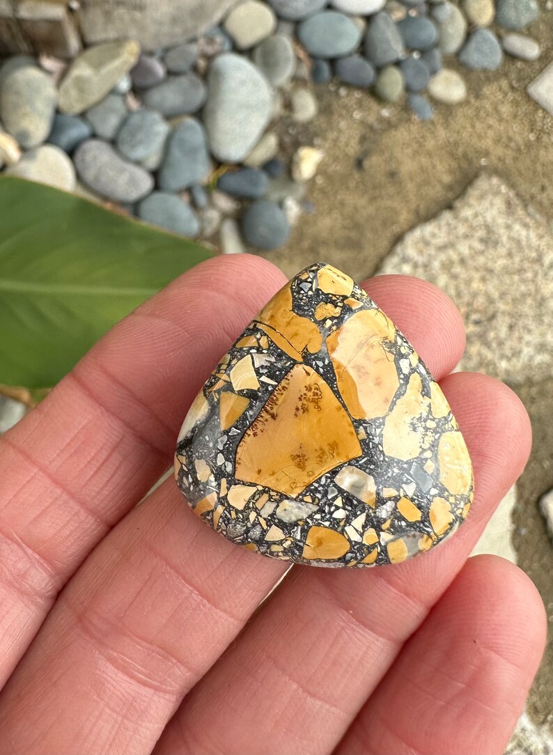 Puede incluir: Una piedra triangular con un patr&oacute;n de mosaico. La piedra presenta una mezcla de fragmentos de color beige, negro y blanco, creando un dise&ntilde;o natural &uacute;nico. La piedra se sostiene en una mano, mostrando su superficie pulida.