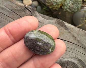 May include: An oval-shaped gemstone with a polished surface. The stone features a blend of green and dark purple hues, with a glossy finish. The gemstone is held in a hand, showcasing its natural colors and textures. The stone is approximately 1.5 inches long.