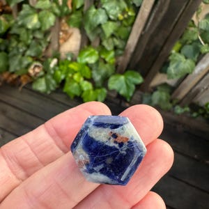 K&ouml;nnte beinhalten: Ein sechseckiger Edelstein mit polierter Oberfl&auml;che. Der Stein weist eine Mischung aus tiefem Blau, Wei&szlig; und Braunt&ouml;nen auf. Die Form ist ein sechsseitiges Polygon, das in einer Hand vor dem Hintergrund von gr&uuml;nem Laub und einem Holzzaun gehalten wird.