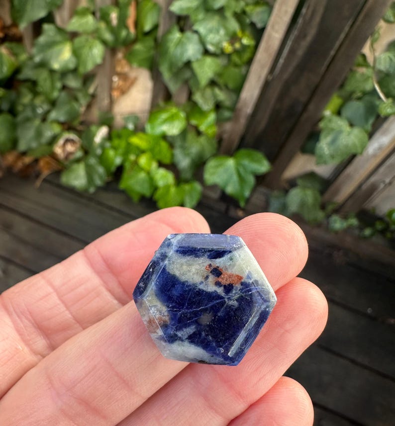 K&ouml;nnte beinhalten: Ein sechseckiger Edelstein mit polierter Oberfl&auml;che. Der Stein weist eine Mischung aus tiefem Blau, Wei&szlig; und Braunt&ouml;nen auf. Die Form ist ein sechsseitiges Polygon, das in einer Hand vor dem Hintergrund von gr&uuml;nem Laub und einem Holzzaun gehalten wird.