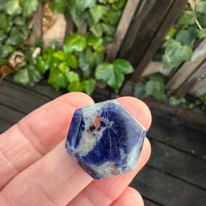 K&ouml;nnte beinhalten: Ein sechseckiger Sodalith-Kristall mit tiefblauer Farbe und wei&szlig;en und braunen Einschl&uuml;ssen. Der polierte Stein wird in einer Hand gehalten, mit einem verschwommenen gr&uuml;nen Hintergrund aus Bl&auml;ttern und Holzlatten.