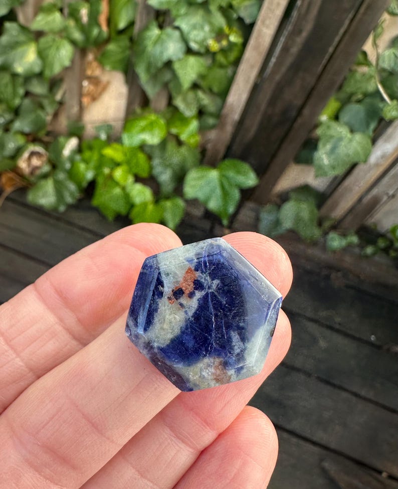 K&ouml;nnte beinhalten: Ein sechseckiger Sodalith-Kristall mit tiefblauer Farbe und wei&szlig;en und braunen Einschl&uuml;ssen. Der polierte Stein wird in einer Hand gehalten, mit einem verschwommenen gr&uuml;nen Hintergrund aus Bl&auml;ttern und Holzlatten.
