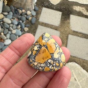 Puede incluir: Una piedra en forma de l&aacute;grima con un patr&oacute;n de mosaico. La piedra presenta una mezcla de fragmentos beige, negros y blancos, creando una apariencia texturizada &uacute;nica. La piedra se sostiene en una mano, mostrando su tama&ntilde;o y forma. Este art&iacute;culo es adecuado para la fabricaci&oacute;n de joyas o como pieza decorativa.