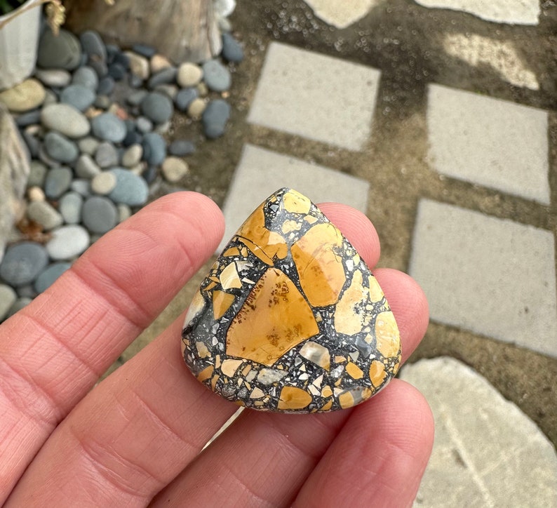 Puede incluir: Una piedra en forma de l&aacute;grima con un patr&oacute;n de mosaico. La piedra presenta una mezcla de fragmentos beige, negros y blancos, creando una apariencia texturizada &uacute;nica. La piedra se sostiene en una mano, mostrando su tama&ntilde;o y forma. Este art&iacute;culo es adecuado para la fabricaci&oacute;n de joyas o como pieza decorativa.