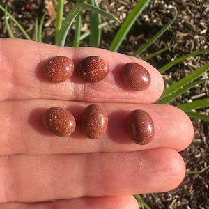May include: Six oval-shaped, reddish-brown gemstones with a sparkling, glittery appearance. The stones are held in a hand, with green plants in the background. The gemstones are likely used for jewelry or crafting.