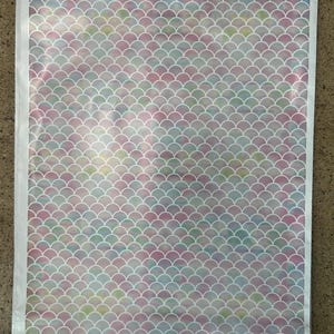May include: A rectangular fabric with a mermaid scale pattern. The scales are in shades of pink, blue, and green, with white outlines. The fabric is framed by a white border.
