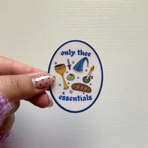 May include: An oval sticker with a white background and a dark blue border. The text 'only thee essentials' is printed in blue. Illustrations include a gold chalice, a small dagger, a blue striped hat, a green apple, a candle, and a loaf of bread.