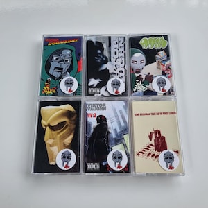 Könnte beinhalten: Sechs Musikkassetten mit Albumcovern des maskierten Charakters MF DOOM. Die Kassetten haben verschiedene Farbschemata, darunter Grün, Schwarz und Beige. Jede Kassette hat einen Aufkleber mit dem MF DOOM Maskenlogo.