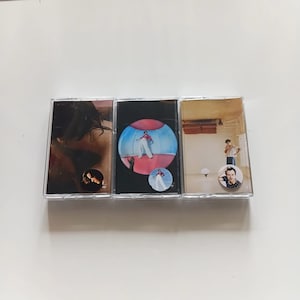 Op de afbeelding: Drie cassettes met albumhoezen. De eerste heeft een donkere afbeelding met een gouden ronde sticker. De tweede heeft een persoon in een blauwe bol. De derde heeft een persoon die in een kamer met een speld staat.