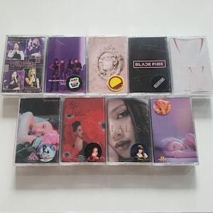 Può includere: Una collezione di dieci musicassette in custodie di plastica trasparente. Le copertine degli album presentano una varietà di colori, tra cui viola, rosso, nero e bianco. Alcuni nastri hanno adesivi. Il testo "BLACKPINK" è visibile su alcuni nastri.
