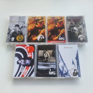 May include: A selection of seven Elliott Smith cassette tapes. The covers showcase diverse artwork, including portraits and abstract designs. The tapes are housed in transparent plastic cases.