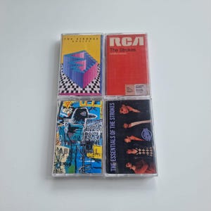 May include: Four cassette tapes with album artwork. The top left tape has a colourful geometric design and the text "The Strokes Angles". The top right tape is red with "RCA The Strokes". The bottom tapes feature abstract art and band photos.