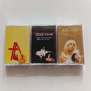 Puede incluir: Tres cintas de cassette con portadas de álbumes de Billie Eilish. Las cintas tienen portadas amarillas, negras y beige. La portada amarilla tiene una estructura roja y la letra "A". La portada negra tiene el texto "Billie Eilish" y "When We All Fall Asleep, Where Do We Go?". La portada beige tiene el texto "Billie Eilish Happier Than Ever".