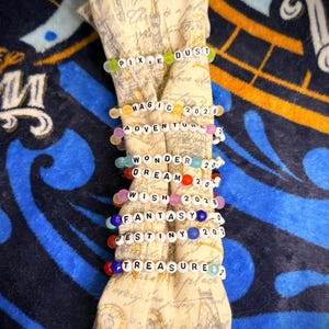 May include: A collection of beaded bracelets with white letter beads spelling out words like "Pixie Dust", "Magic", "Adventure", "Wonder", "Dream", "Wish", "Fantasy", "Destiny", and "Treasure". The bracelets are colorful and strung on elastic.