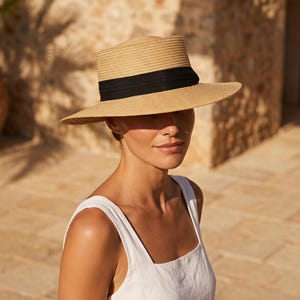 May include: A tan straw fedora hat featuring a structured crown and a wide, flat brim. The hat is accented with a solid black fabric ribbon band wrapped around the base of the crown, providing a distinct contrast against the natural woven texture.