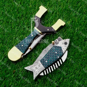May include: A slingshot and a folding knife with a fish design are shown on green grass. The slingshot has a black and gold frame with a green and black patterned handle. The knife is fish-shaped with a green and black honeycomb pattern.