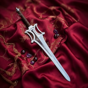 May include: A silver-toned dagger with a brown wrapped handle and a stylized crossguard. The blade features a central ridge and pointed tip. Several small, smooth, dark-colored stones are scattered nearby, and a gold-toned chain is visible.