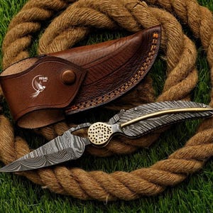 May include: A folding knife with a Damascus steel blade and a handle shaped like a feather, featuring a brass pivot point with a perforated design. The knife is accompanied by a brown leather sheath embossed with a bird logo and the text REGAL.