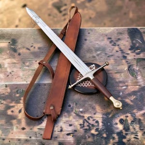 Puede incluir: Una espada de acero con acanaladura central, guarda recta y empuñadura forrada en cuero marrón. La espada presenta un pomo de latón y se acompaña de una vaina de cuero marrón a juego con una correa de hombro ajustable integrada.