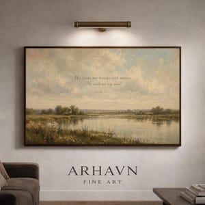 May include: A framed landscape painting depicts a serene waterscape under a cloudy sky. The artwork features a quote: "He leads me beside still waters, He restores my soul." The bottom of the frame displays the text "ARHAVN FINE ART".