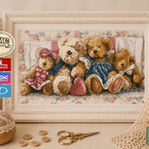 May include: A framed cross-stitch embroidery featuring four teddy bears sitting together on a white pillow. The bears are brown and tan, wearing small outfits including a pink dress and a blue dress. A red heart sits in front of the bears.