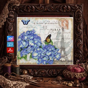 May include: Cross-stitch artwork in a dark wooden frame. The artwork features blue hydrangeas, a butterfly, and the Eiffel Tower. The words "Carte Postale" and "Paris" are visible. The frame is surrounded by sewing notions.